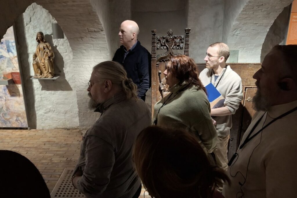 Grupa zwiedzających zgromadzona w muzealnej sali z ceglanym sklepieniem, słuchająca oprowadzania i oglądająca rzeźby oraz eksponaty (kliknięcie powiększy zdjęcie)