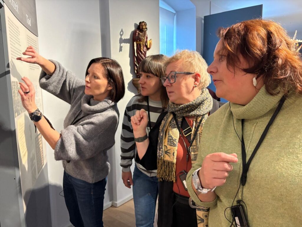 Uczestnicy oprowadzania stoją przy planszy informacyjnej, jedna z osób wskazuje fragment tekstu na ścianie ekspozycji (kliknięcie powiększy zdjęcie)