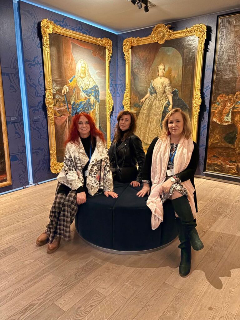 Trzy osoby siedzą na ławce w sali muzealnej, za nimi dwa duże obrazy w bogato zdobionych złoconych ramach (kliknięcie powiększy zdjęcie)