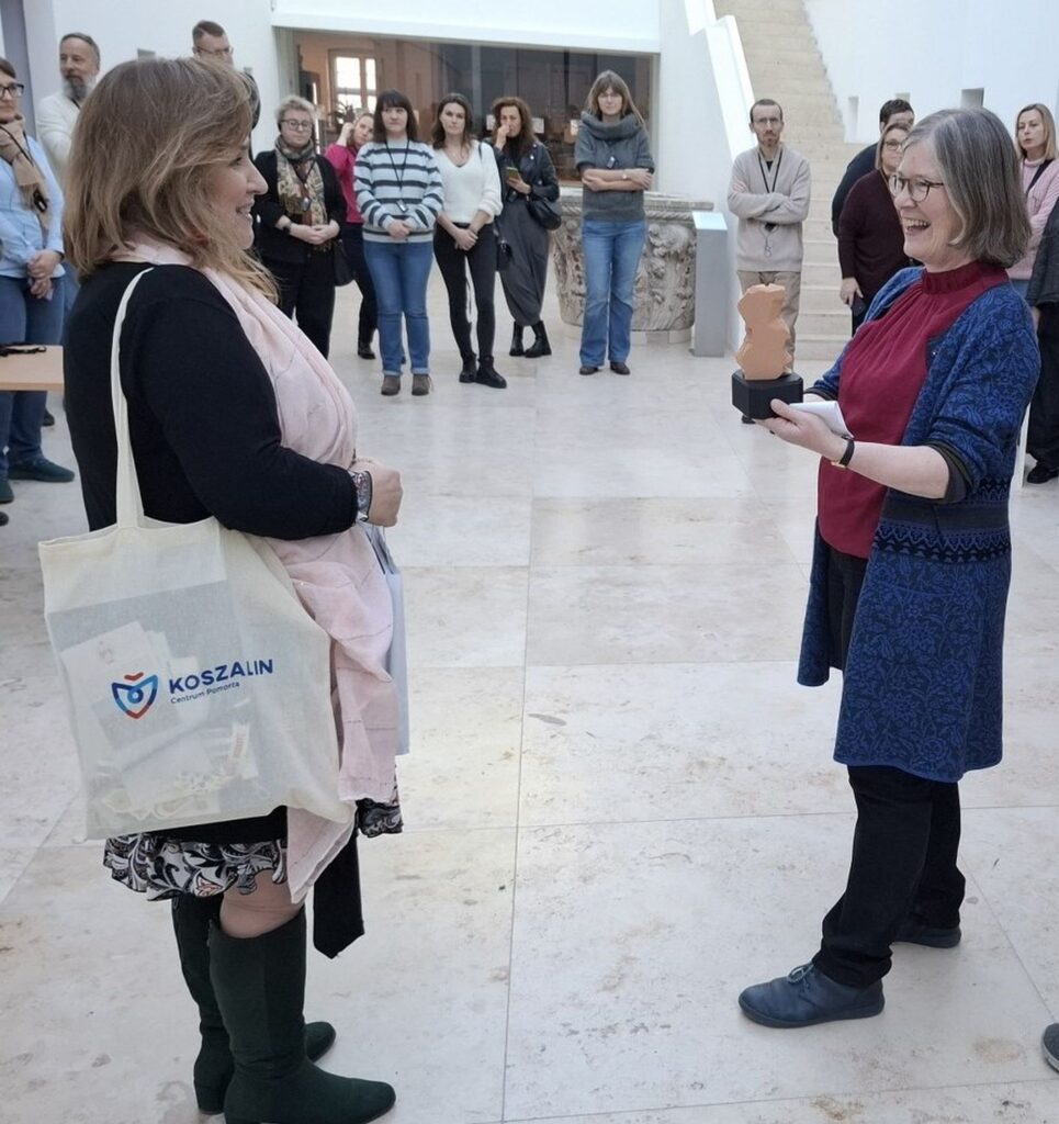 Uczestnicy oprowadzania stojący w sali i słuchają osoby prowadzącej oraz oglądający eksponaty (kliknięcie powiększy zdjęcie)