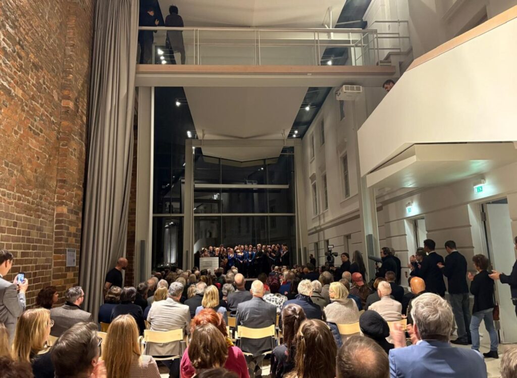 Wnętrze nowoczesnego muzeum: liczna publiczność siedząca i stojąca podczas wydarzenia, wysoka przestrzeń z antresolą i ceglaną ścianą (kliknięcie powiększy zdjęcie)