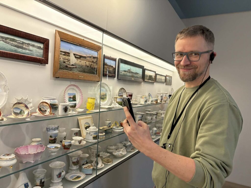 Zwiedzający pozuje do zdjęcia - przy okazji fotografuje gablotę z porcelaną i naczyniami, ustawioną wzdłuż ściany sali muzealnej (kliknięcie powiększy zdjęcie)