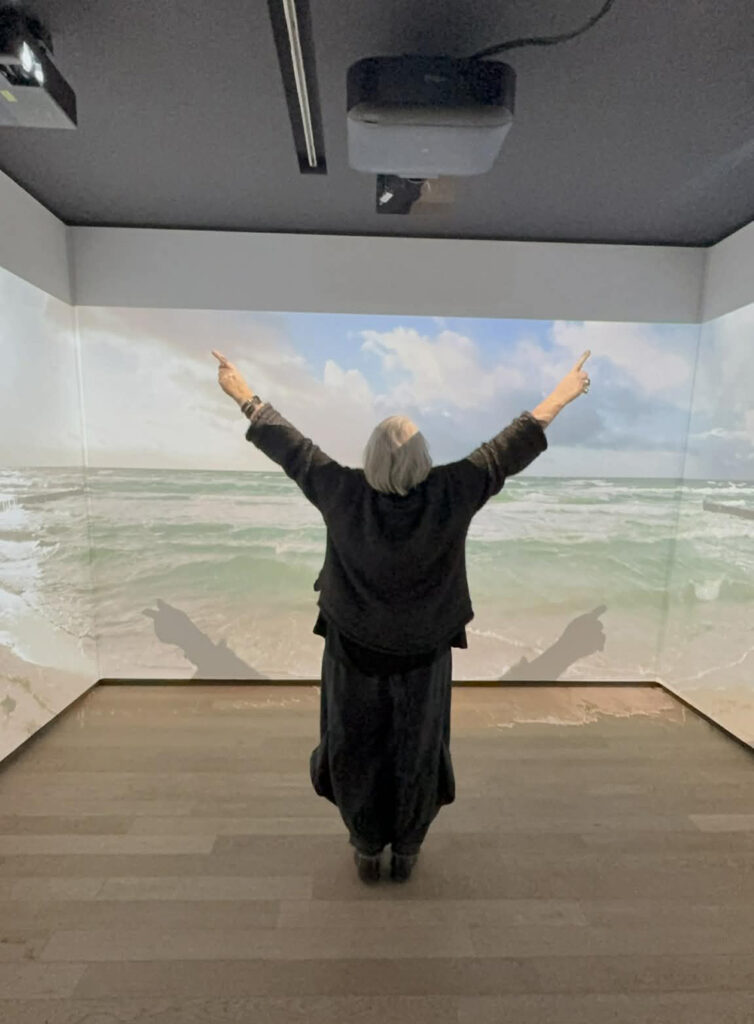 Osoba stoi w sali immersyjnej przed panoramiczną projekcją morza, z uniesionymi rękami skierowanymi ku górze (kliknięcie powiększy zdjęcie)