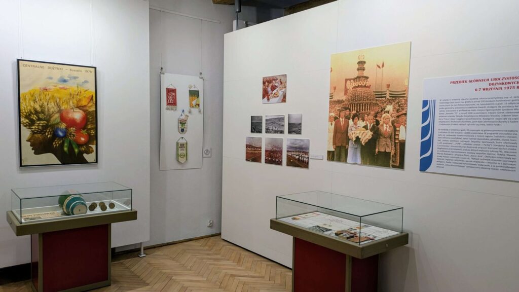 Fragment wystawy dożynkowej z gablotą muzealną oraz planszami i fotografiami dokumentującymi tradycje święta plonów, prezentowanymi na białych ścianach sali (kliknięcie powiększy zdjęcie).
