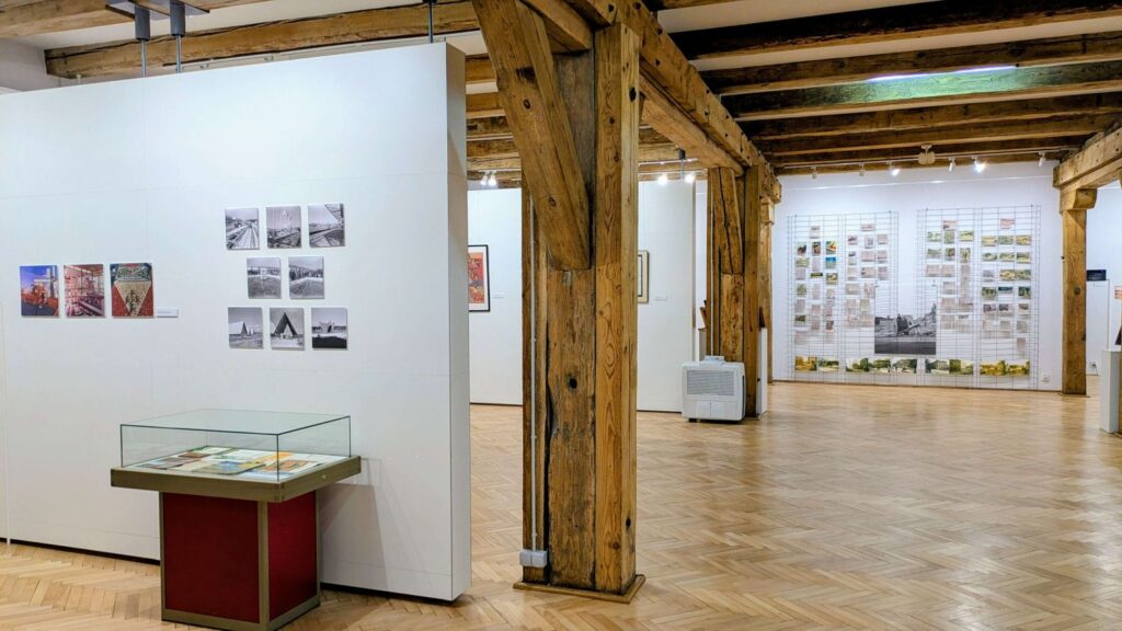 Widok sali wystawowej z planszami fotograficznymi i gablotą ekspozycyjną, w tle dalsza część ekspozycji dożynkowej (kliknięcie powiększy zdjęcie).