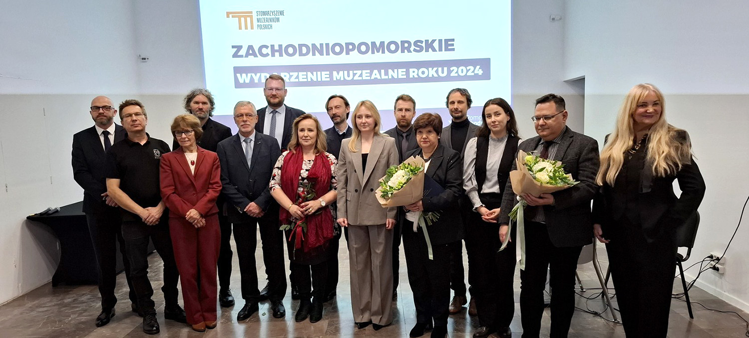 Kilkunastoosobowa grupa ludzi stoi przed wyświetlanym na ścianie napisem "Zachodniopomorskie Wydarzenie Muzealne Roku 2024". Dwie z tych osób trzymają w dłoni bukiety kwiatów.