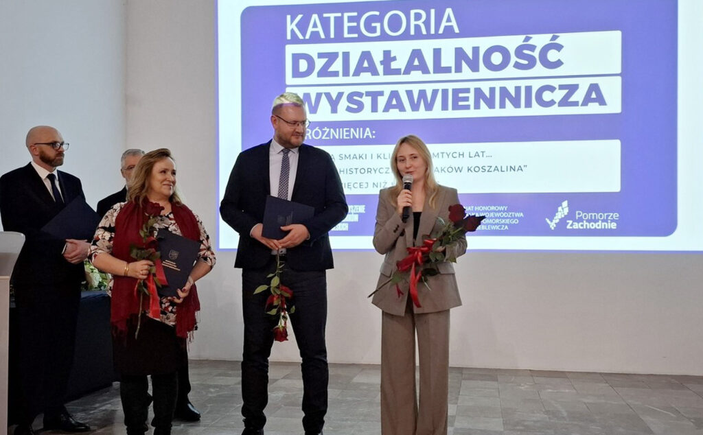 Grupa osób stoi przed ekranem, na którym wyświetla się slajd "Kategoria Działalność Wystawiennicza". Jedna osoba przemawia do mikrofonu (kliknięcie powiększy zdjęcie)