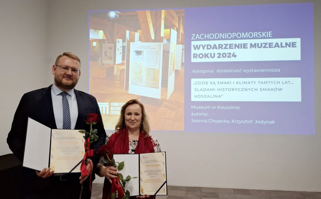 Dwie osoby trzymające dyplomy i czerwone róże. Stoją przed ekranem, na którym wyświetla się slajd "Zachodniopomorskie Wydarzenie Muzealne Roku 2024" (kliknięcie powiększy zdjęcie)