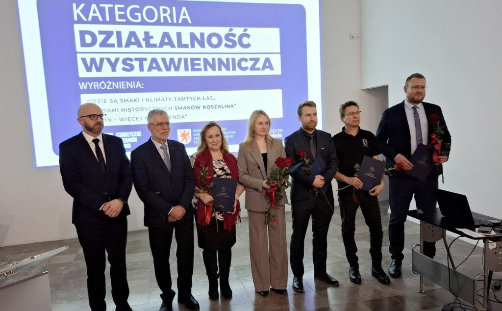 Laureaci i organizatorzy stojący wspólnie przed ekranem, na którym wyświetla się slajd "Kategoria Działalność Wystawiennicza". Osoby trzymają dyplomy i kwiaty (kliknięcie powiększy zdjęcie)