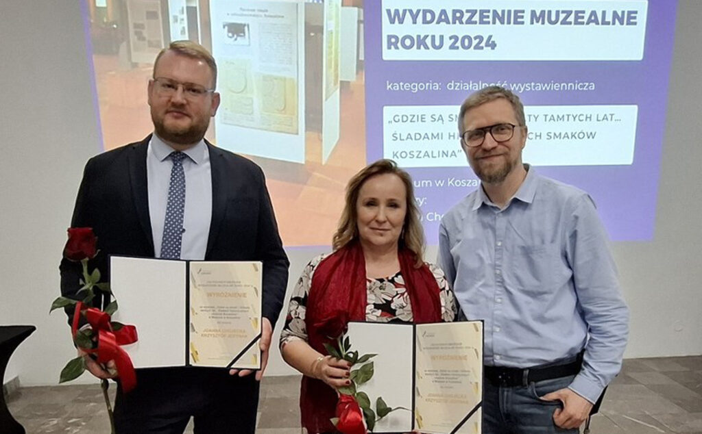 Trzy osoby stoją przed ekranem z prezentacją  „Wydarzenie Muzealne Roku 2024” i trzymają dyplomy oraz czerwone róże (kliknięcie powiększy zdjęcie)