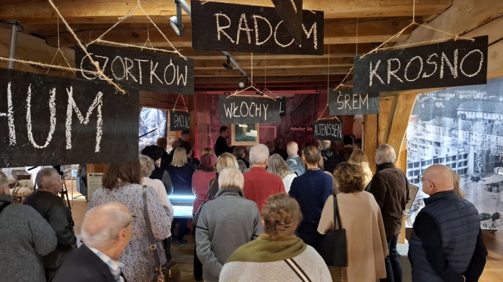 Publiczność zgromadzona w przestrzeni wystawowej z drewnianymi stropami; nad głowami wiszą tablice z nazwami miejscowości, w tle ekspozycja muzealna (kliknięcie powiększy zdjęcie).
