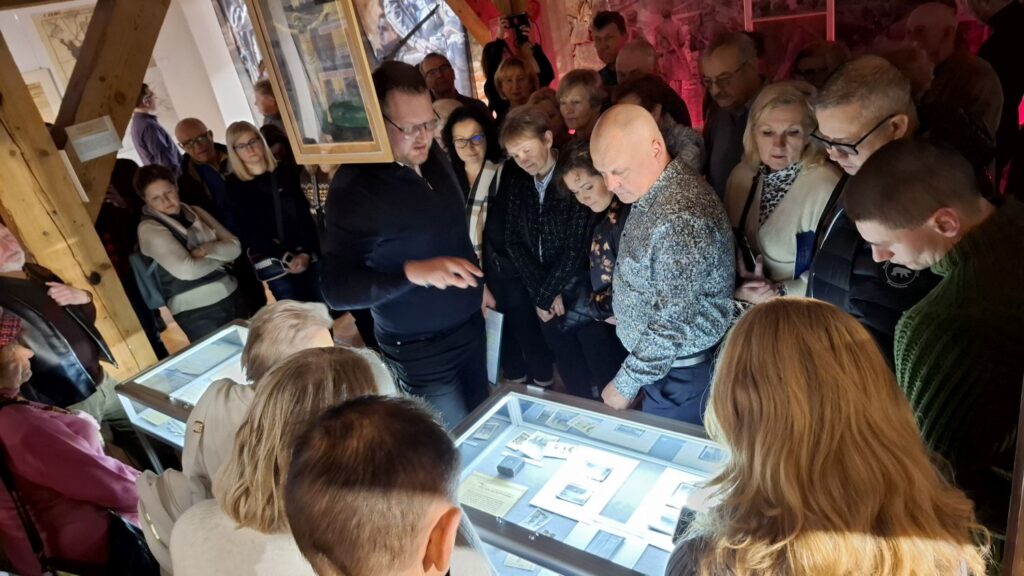 Grupa zwiedzających stojąca wokół podświetlonej gabloty z materiałami archiwalnymi; uczestnicy uważnie oglądają prezentowane dokumenty (kliknięcie powiększy zdjęcie).