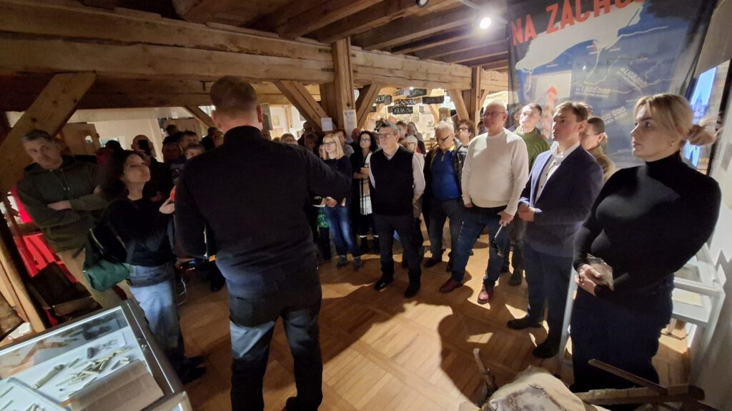 Grupa uczestników stojąca w sali wystawowej, słuchająca oprowadzania wśród eksponatów i plansz (kliknięcie powiększy zdjęcie).
