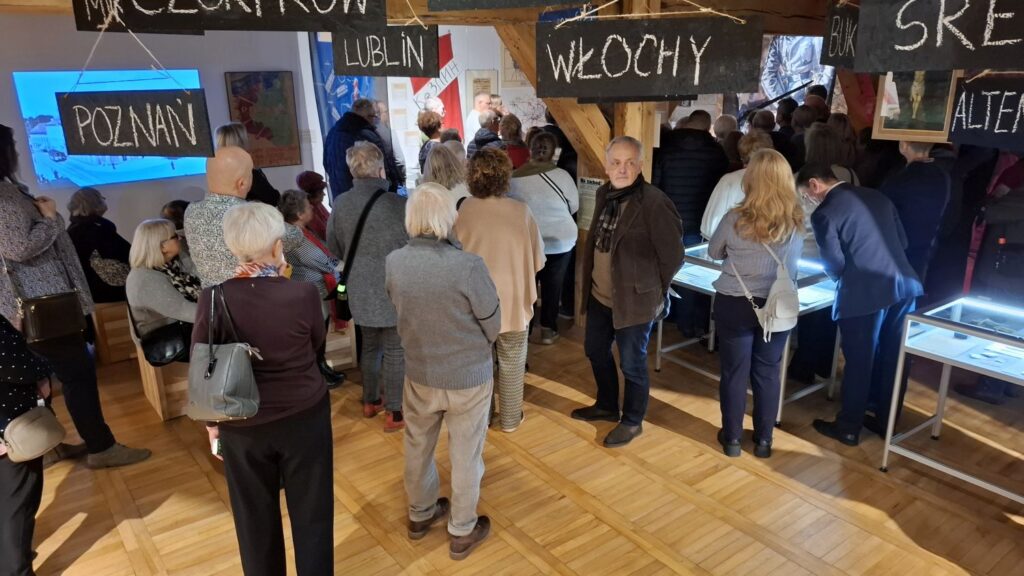 Tłum odwiedzających zgromadzony przy gablotach w sali wystawowej z tablicami nazw miejscowości nad ekspozycją (kliknięcie powiększy zdjęcie).