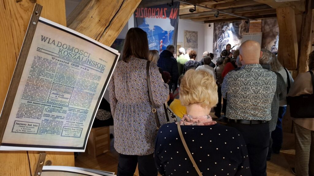 Uczestnicy wydarzenia stojący przed planszą z archiwalnym artykułem prasowym w przestrzeni wystawy (kliknięcie powiększy zdjęcie).