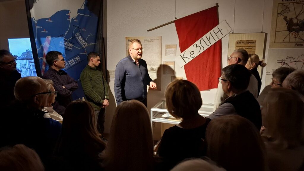 Grupa uczestników zgromadzona wokół prowadzącego, który opowiada o ekspozycji; w tle plansze i historyczna flaga z napisem „Kozienice” (kliknięcie powiększy zdjęcie).