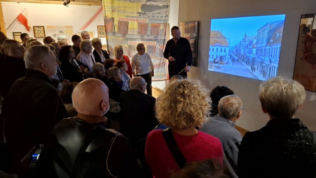 Uczestnicy wydarzenia stojący przed ekranem z archiwalną fotografią ulicy, słuchający komentarza prowadzącego (kliknięcie powiększy zdjęcie).