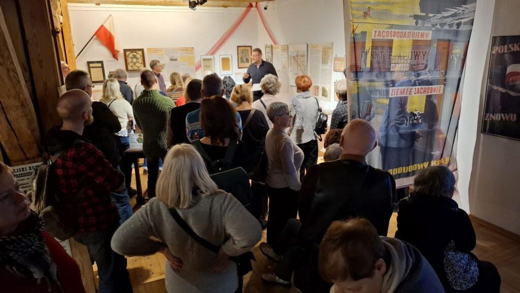 Liczna publiczność zebrana w sali wystawowej podczas oprowadzania, w otoczeniu plansz i materiałów archiwalnych (kliknięcie powiększy zdjęcie).