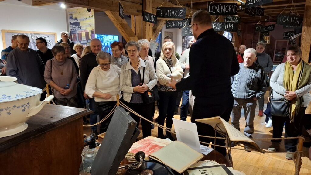 Zwiedzający zgromadzeni wokół przewodnika w przestrzeni ekspozycji, na pierwszym planie historyczne przedmioty i dokumenty (kliknięcie powiększy zdjęcie).