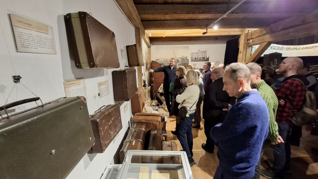 Zwiedzający oglądający eksponaty umieszczone na ścianach i w gablotach w sali muzealnej z drewnianą konstrukcją (kliknięcie powiększy zdjęcie).