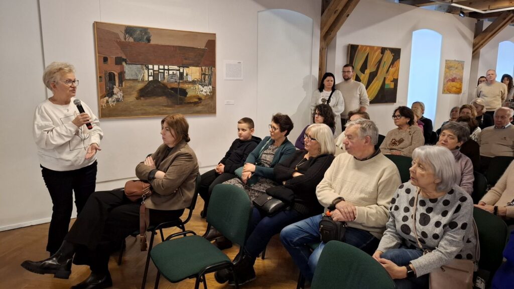 Kobieta stojąca z mikrofonem zabiera głos wśród siedzącej publiczności w muzeum (kliknięcie powiększy zdjęcie)