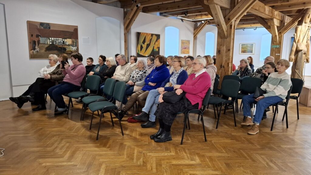Uczestnicy wydarzenia siedzący w rzędach krzeseł w sali muzealnej, na ścianach widoczne obrazy (kliknięcie powiększy zdjęcie)