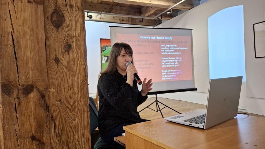 Prowadząca wykład kobieta mówi do mikrofonu przy stole z laptopem, za nią ekran z prezentacją w sali muzealnej (kliknięcie powiększy zdjęcie)