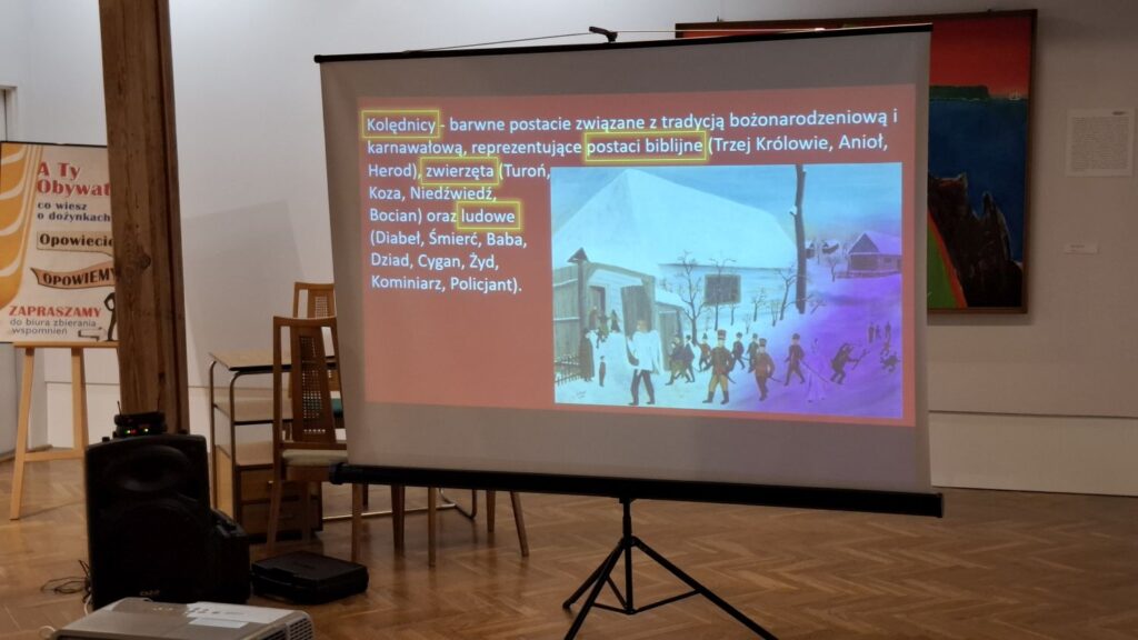 Ekran projekcyjny z planszą prezentacji oraz fragment sali muzealnej z krzesłami dla publiczności (kliknięcie powiększy zdjęcie)