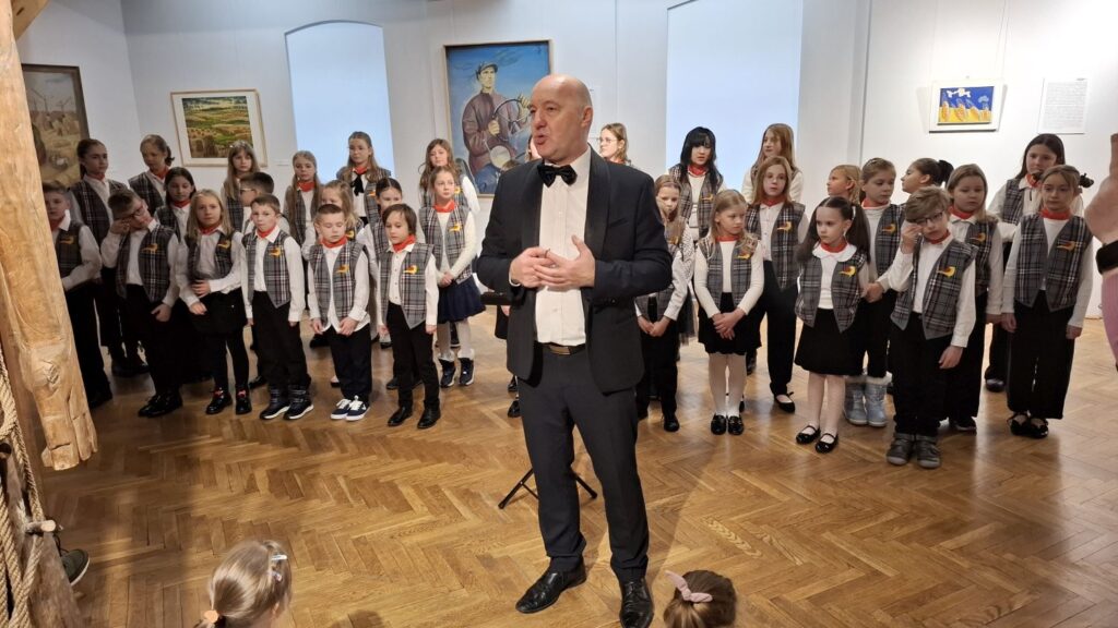 Dyrygent stojący przed dziecięcym chórem przemawia do publiczności zgromadzonej w muzeum (kliknięcie powiększy zdjęcie)