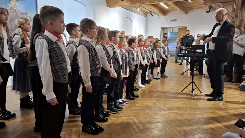 Dyrygent prowadzący dziecięcy chór ustawiony w rzędzie, w tle widoczna publiczność (kliknięcie powiększy zdjęcie)