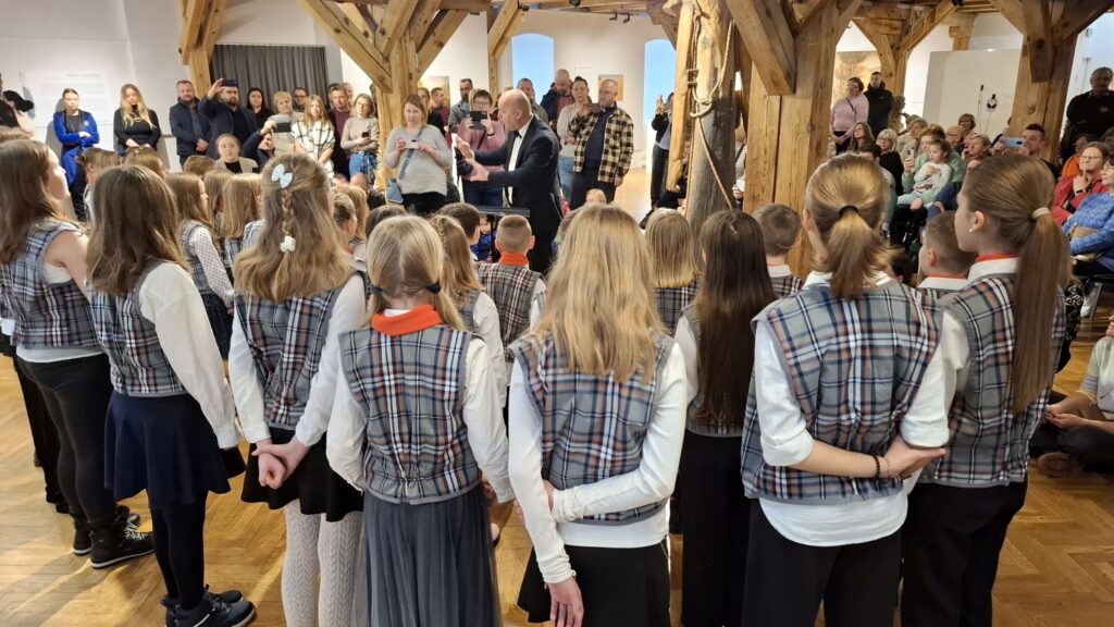 Widok od tyłu na dziecięcy chór stojący przed zgromadzoną publicznością w sali muzealnej (kliknięcie powiększy zdjęcie)