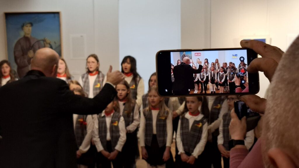 Osoba nagrywająca występ dziecięcego chóru smartfonem, w tle dyrygent i śpiewające dzieci (kliknięcie powiększy zdjęcie)