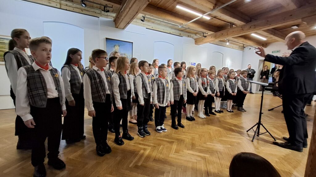 Dziecięcy chór ustawiony w rzędzie śpiewa w sali muzealnej, przed nimi dyrygent prowadzący występ (kliknięcie powiększy zdjęcie)