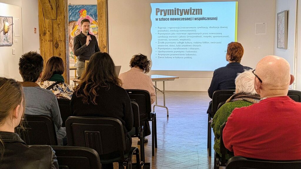 Prelegent stojący przed publicznością na sali wystawowej, obok niego slajd z tytułem „Prymitywizm w sztuce nowoczesnej i współczesnej” wyświetlany na ekranie (kliknięcie powiększy zdjęcie).