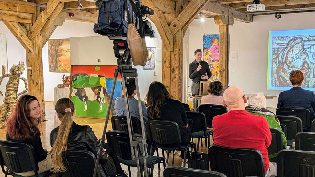 Wnętrze sali wystawowej z publicznością siedzącą na krzesłach podczas wykładu; na pierwszym planie kamera na statywie, w tle obrazy na ścianach (kliknięcie powiększy zdjęcie).