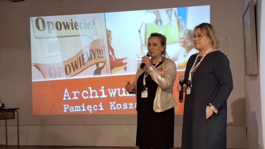 Dwie osoby stojące przed ekranem z prezentacją „Archiwum Pamięci Koszalina”, jedna z nich przemawia do publiczności (kliknięcie powiększy zdjęcie)