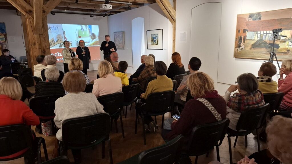 Publiczność siedząca na sali wystawowej podczas spotkania, w tle przemawiają prelegenci (kliknięcie powiększy zdjęcie)