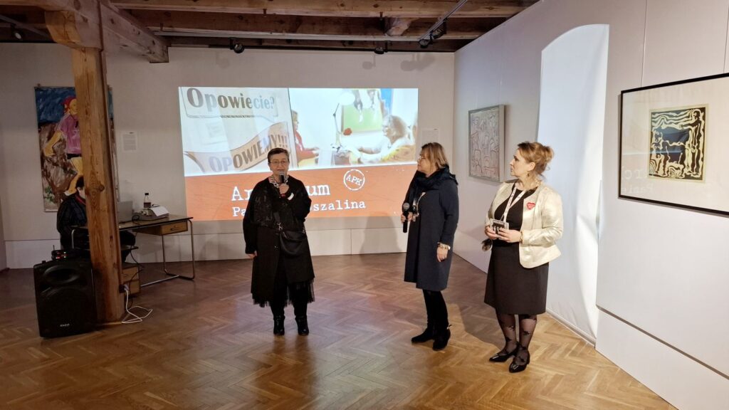 Trzy osoby stojące na sali wystawowej, jedna z nich trzyma w ręku mikrofon i przemawia do publiczności (kliknięcie powiększy zdjęcie)