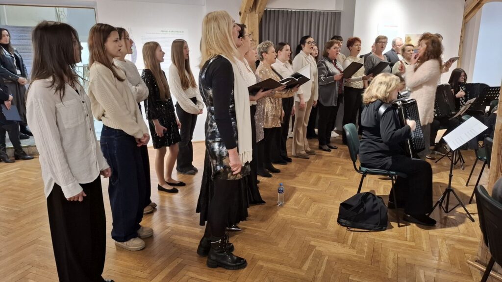Chór ustawiony w rzędzie w sali wystawowej, śpiewacy trzymający nuty i wykonujący utwory podczas koncertu (kliknięcie powiększy zdjęcie)