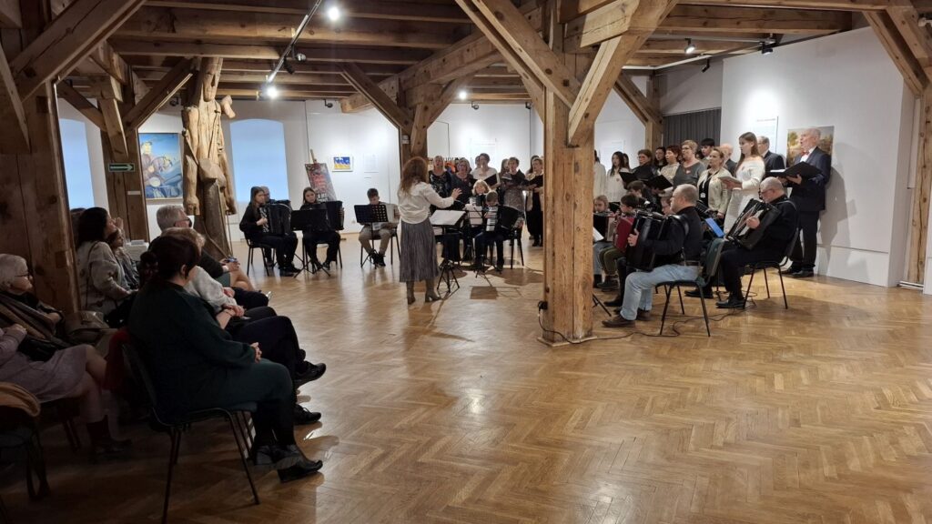 Widok ogólny sali wystawowej podczas koncertu, publiczność siedząca wokół muzyków i chóru w przestrzeni galerii (kliknięcie powiększy zdjęcie)