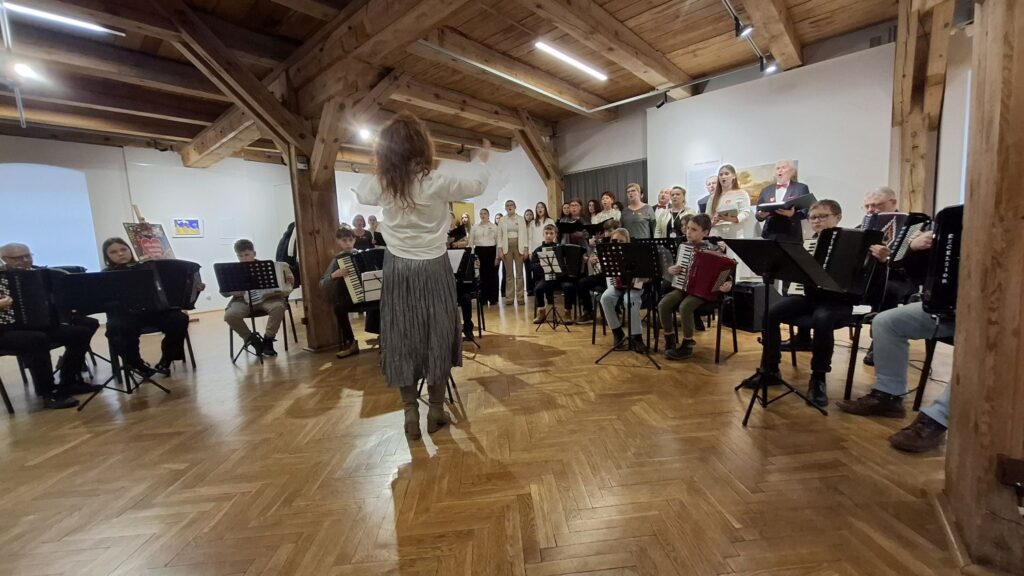 Dyrygentka stojąca na środku sali wystawowej i prowadząca zespół muzyków oraz chór podczas koncertu (kliknięcie powiększy zdjęcie