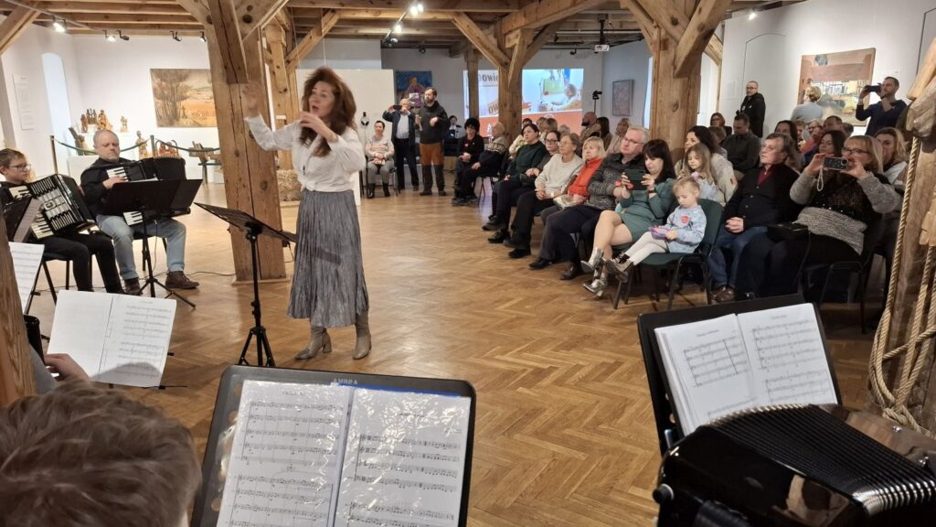 Dyrygentka prowadząca koncert, w tle chór i publiczność siedząca w sali wystawowej (kliknięcie powiększy zdjęcie