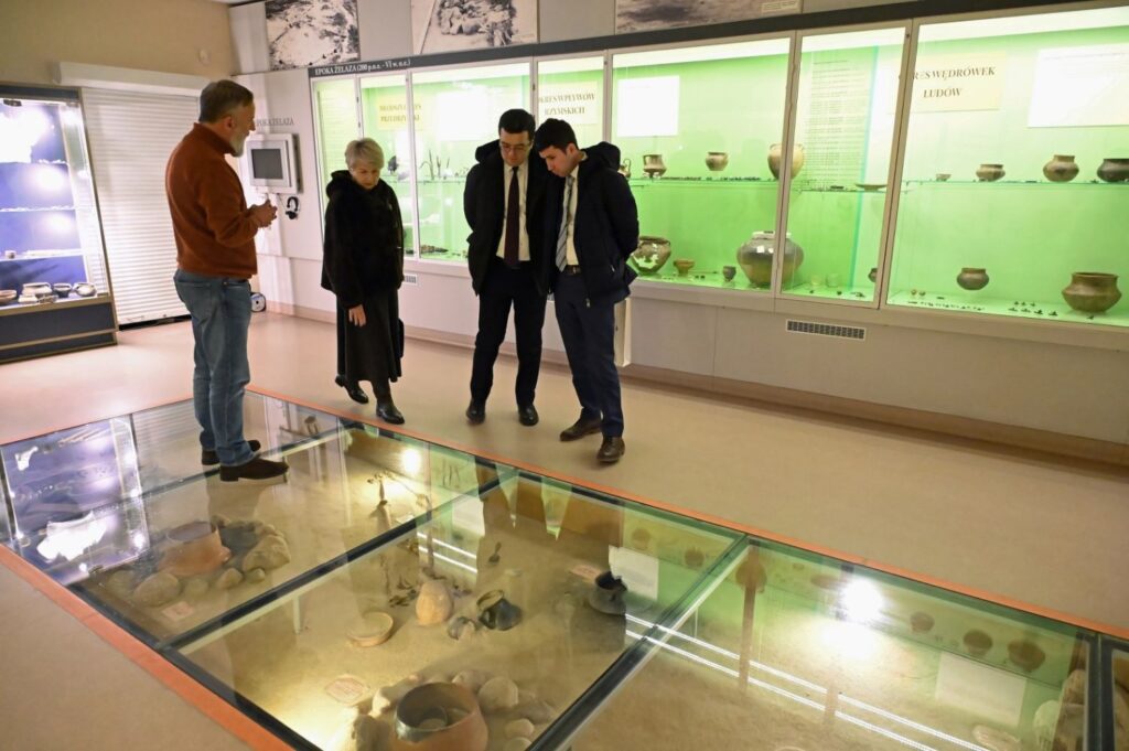 Uczestnicy zwiedzania oglądający eksponaty muzealne umieszczone w przeszklonych gablotach podłogowych (kliknięcie powiększy zdjęcie).
