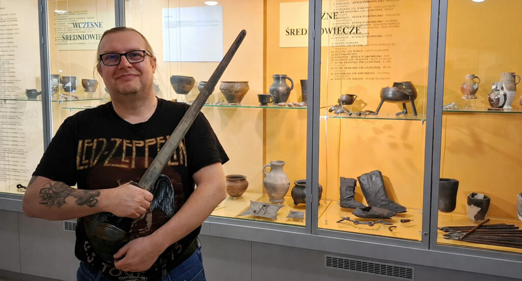 Mężczyzna trzyma replikę wczesnośredniowiecznego miecza, stojąc przed gablotą muzealną z zabytkowymi naczyniami, narzędziami i obuwiem z okresu średniowiecza (kliknięcie powiększy zdjęcie).