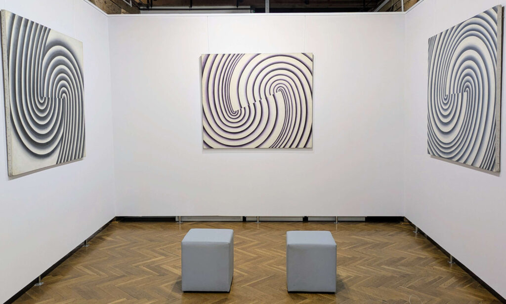 Ścianka wystawowa z trzema obrazami przedstawiającymi spiralne wzory w czerni i bieli, na środku dwa szare siedziska (kliknięcie powiększy zdjęcie).