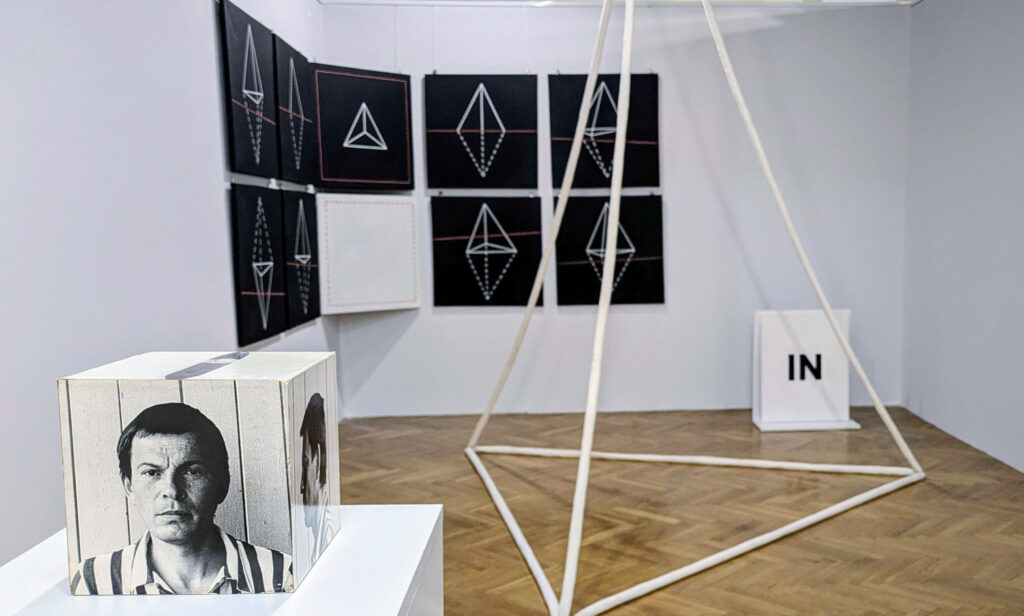 Fragment ekspozycji sztuki współczesnej z instalacją przestrzenną z białych elementów, czarno-białymi grafikami geometrycznymi na ścianie oraz sześciennym obiektem z portretem (kliknięcie powiększy zdjęcie).