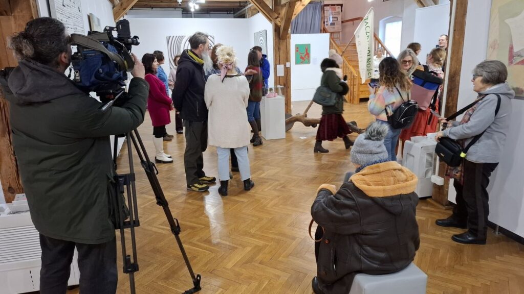 Uczestnicy otwarcia wystawy zgromadzeni w sali ekspozycyjnej; w tle widoczna ekipa telewizyjna z kamerą rejestrująca wydarzenie oraz oglądane prace artystyczne (kliknięcie powiększy zdjęcie)