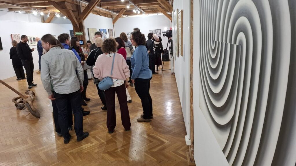 Zwiedzający oglądający prace podczas wernisażu; na pierwszym planie fragment abstrakcyjnego obrazu o koncentrycznych kręgach, w tle osoby poruszające się po sali wystawowej (kliknięcie powiększy zdjęcie)