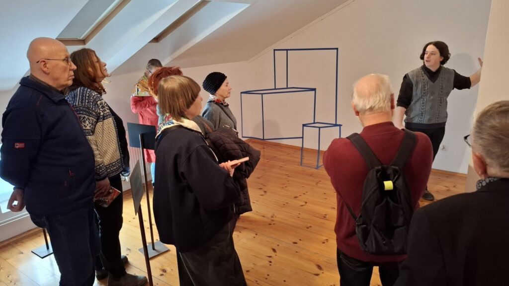 Oprowadzanie po wystawie – osoba prowadząca wskazuje na geometryczną instalację przestrzenną, a zgromadzeni goście słuchają wyjaśnień w sali ekspozycyjnej (kliknięcie powiększy zdjęcie)