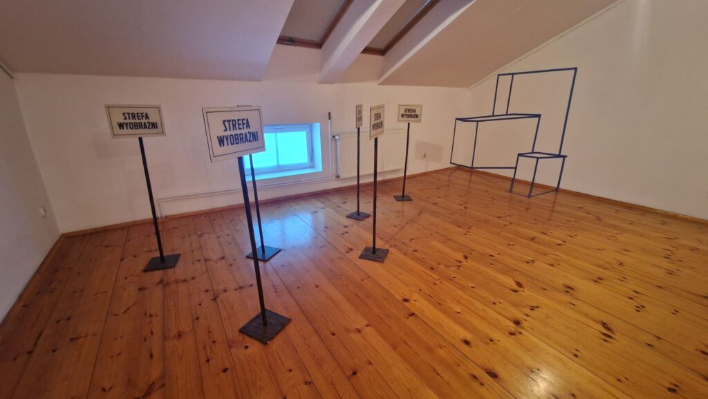 Widok sali wystawowej z drewnianą podłogą; na środku ustawione są stojaki z tabliczkami o treści "Strefa wyobraźni" oraz schematyczna, liniowa instalacja ścienna (kliknięcie powiększy zdjęcie)